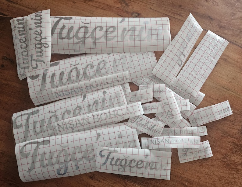May include: A collection of silver vinyl decals on white transfer paper with a red grid pattern. The decals feature the text Tuğ&ccedil;e'nin and NİŞAN BOH&Ccedil;ASI in a cursive script font, designed for application onto various surfaces as custom labels.
