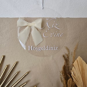 May include: A clear round acrylic sign with a white ribbon bow and the text "Kiz Evine Hosgeldiniz" which translates to "Welcome to the Girl's House".