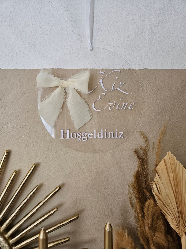 May include: A clear round acrylic sign with a white ribbon bow and the text "Kiz Evine Hosgeldiniz" which translates to "Welcome to the Girl's House".