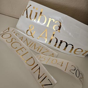 May include: Custom gold foil adhesive vinyl decals on white backing paper. The text reads K&uuml;bra & Ahmet, HOŞGELDİNİZ, İLK ANIMIZA, and 05/04/2025. These metallic lettering stickers feature a serif font and are designed for application on smooth surfaces.