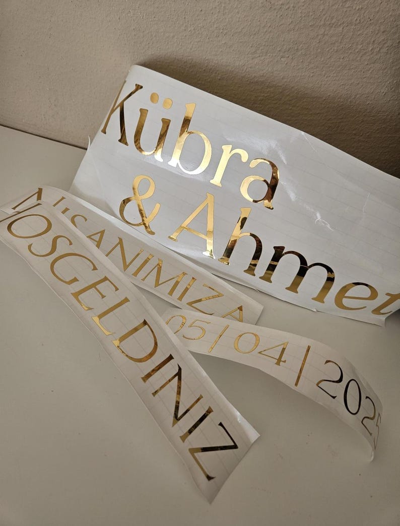 May include: Custom gold foil adhesive vinyl decals on white backing paper. The text reads K&uuml;bra & Ahmet, HOŞGELDİNİZ, İLK ANIMIZA, and 05/04/2025. These metallic lettering stickers feature a serif font and are designed for application on smooth surfaces.