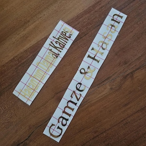 May include: Two rectangular adhesive vinyl decals with gold metallic lettering on a white backing paper. One decal features the text "Hasan'ın Damat Kahvesi" and the other displays the names "Gamze & Hasan" in a cursive script font.