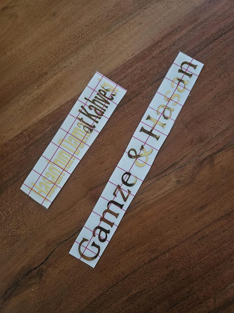 May include: Two rectangular adhesive vinyl decals with gold metallic lettering on a white backing paper. One decal features the text "Hasan'ın Damat Kahvesi" and the other displays the names "Gamze & Hasan" in a cursive script font.