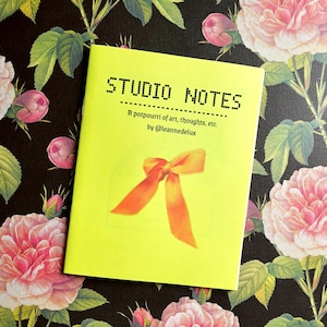 May include: A bright yellow notebook titled "STUDIO NOTES" with the text "A potpourri of art, thoughts, etc. by @leannedelux" and an orange bow illustration. The notebook rests on a floral patterned background with pink roses and green leaves.