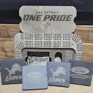 Può includere: Quattro sottobicchieri in ardesia a tema Detroit Lions. I sottobicchieri presentano il logo della squadra, le parole "Lions" e "Ford Field". I sottobicchieri sono disposti a forma di quadrato. Un cartello in legno con le parole "One Detroit One Pride" è sullo sfondo.