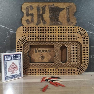 May include: A wooden game board with a Minnesota outline and the word "SKOL" on the lid. The board is designed for a game of cribbage and features a small compartment for holding playing cards. The board is accompanied by a deck of Maverick playing cards and red and white wooden pegs.