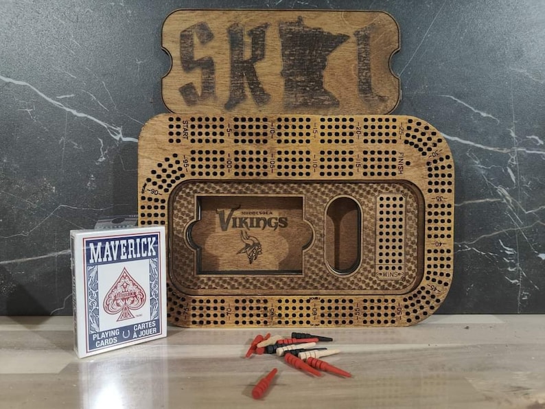May include: A wooden game board with a Minnesota outline and the word "SKOL" on the lid. The board is designed for a game of cribbage and features a small compartment for holding playing cards. The board is accompanied by a deck of Maverick playing cards and red and white wooden pegs.
