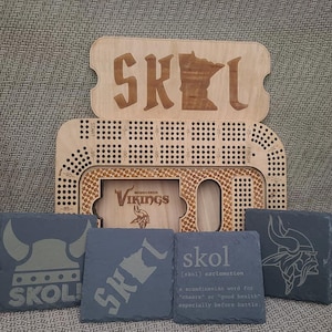 May include: A set of four black slate coasters with different designs. One coaster features a Viking helmet, another has the word "SKOL!" with a Minnesota outline, another has the word "skol" with a definition, and the last one has a Minnesota Vikings logo.