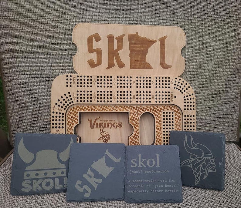 May include: A set of four black slate coasters with different designs. One coaster features a Viking helmet, another has the word "SKOL!" with a Minnesota outline, another has the word "skol" with a definition, and the last one has a Minnesota Vikings logo.