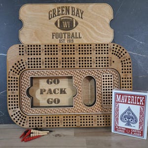 May include: A wooden game board with a green and white football design. The board is labeled "Green Bay WI Football Est. 1919". The board has a grid of holes and a "Go Pack Go" inscription.