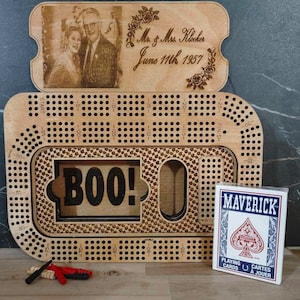 Op de afbeelding: Een houten spelbord met een "BOO!" -uitsparing en een klein fotolijstje met een trouwfoto. Het lijstje heeft de tekst "Mr. & Mrs. Klaker June 11th 1957". Het spelbord heeft een raster van gaten en een baan rond de buitenkant. Een kaartspel bevindt zich in de rechterbenedenhoek.