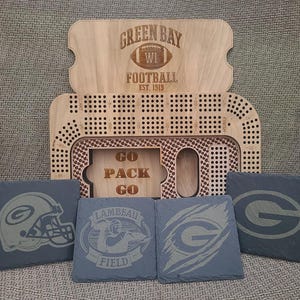 Op de afbeelding: Een houten spelbord met een Green Bay Packers-thema. Het bord is gemaakt van hout en heeft een groen-gouden kleurenschema. Het bord is omringd door vier zwarte leisteenonderzetters met verschillende Green Bay Packers-logo's. De onderzetters zijn gemaakt van leisteen en hebben een zwart-groen kleurenschema. De onderzetters zijn versierd met het Green Bay Packers-logo, de woorden "Lambeau Field" en de woorden "Go Pack Go".