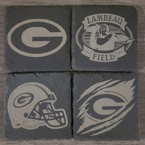 May include: Set of four black slate coasters with different Green Bay Packers logos. The logos include the team's iconic "G", a helmet, a "G" with a claw mark, and a logo with the words "Lambeau Field" and the years "1957" and "1997".