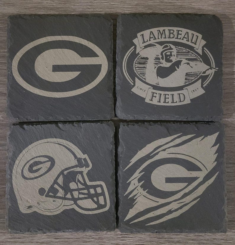May include: Set of four black slate coasters with different Green Bay Packers logos. The logos include the team's iconic "G", a helmet, a "G" with a claw mark, and a logo with the words "Lambeau Field" and the years "1957" and "1997".