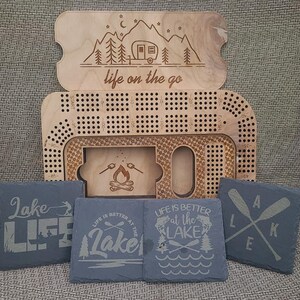 May include: A wooden game board with a camping theme. The board is engraved with a camper, trees, stars, and the words "life on the go". The board is accompanied by four black slate coasters with various lake-themed designs and text, including "Lake Life", "Life is Better at the Lake", and "Lake".