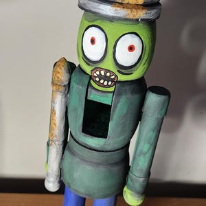 May include: A hand-painted wooden zombie nutcracker. The figure has a green head, a crown, and blue trousers. The nutcracker is painted with a distressed finish, giving it a vintage appearance. The height is approximately 20 cm.