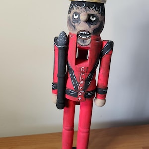 May include: A wooden nutcracker figure wearing a gold crown, a red jacket with black accents, and red trousers. The figure has a painted face with dark shading around the eyes, a wide open mouth showing teeth, and holds a black staff in one hand.
