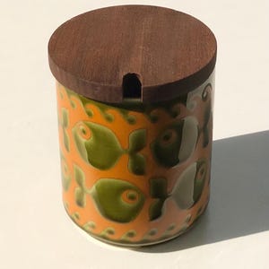 May include: A ceramic jar with a wooden lid. The jar is orange with a repeating pattern of green fish. The lid is brown and has a small opening. A decorative kitchen item.