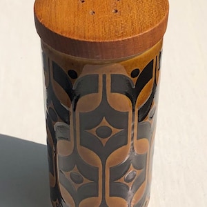 May include: A cylindrical salt or pepper shaker featuring a brown wooden lid with five small holes. The body of the shaker is made of ceramic with a glossy finish, decorated with a repeating geometric pattern in shades of dark brown and tan.