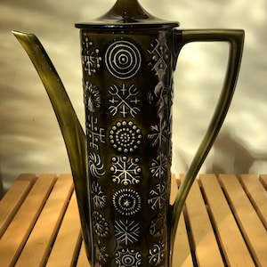 May include: A tall, cylindrical olive green ceramic coffee pot with a matching lid featuring a pointed finial. The exterior surface is decorated with a vertical pattern of white, etched geometric symbols, including circles, stars, and cross-like shapes.