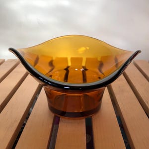 May include: An amber-coloured glass bowl with a distinctive, wavy rim and a dark brown trim. The bowl sits on a short, cylindrical base with a matching dark brown band. The design is reminiscent of mid-century modern decor.
