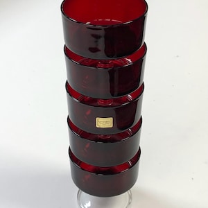 May include: A tower of six ruby red glass bowls, each with a rounded shape and a clear, circular base. The bowls are stacked on top of each other, creating a tower-like structure. A small gold label is visible on one of the bowls.