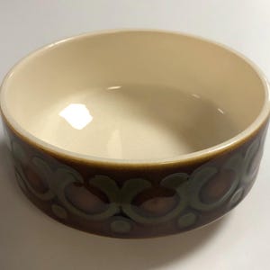 May include: A ceramic bowl with a cream interior and a decorative band around the exterior. The band features a repeating pattern of brown, green, and maroon floral shapes. The bowl has a simple, rounded shape.