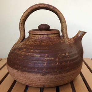 May include: A handcrafted, rustic ceramic teapot with a large arched handle and a small spout. The teapot is primarily brown with lighter brown and tan accents, and a small, round lid. The teapot sits on a wooden surface.