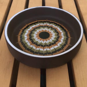 May include: A brown ceramic bowl with a white rim. The interior has a concentric pattern of green, orange, black, and white radiating from a central black circle. The bowl is on a wooden surface.