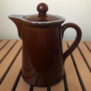May include: A brown ceramic creamer with a lid and a handle. The creamer has a white rim around the top.