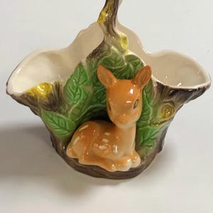 May include: A ceramic planter shaped like a tree stump with a deer figurine and green leaves. The planter has a handle shaped like a branch.