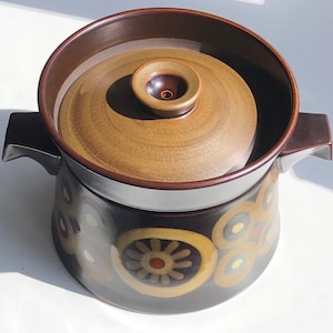 May include: A vintage ceramic casserole dish in shades of brown. The dish features a lid and two handles. The exterior is dark brown with a floral pattern in yellow, orange, and green. The lid and interior are a lighter brown.