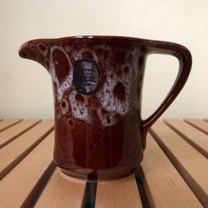 May include: A brown ceramic jug with a white glaze and a label that reads "Shawmer Pottery, Craven, Yorkshire".