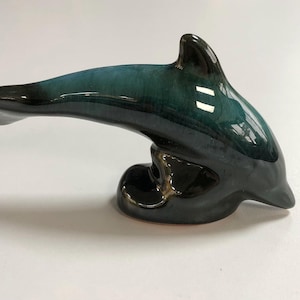 May include: A ceramic dolphin ornament with a glossy finish. The dolphin is teal and black, with a curved body and a streamlined design. The ornament is a decorative item for the home.