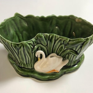 May include: A green ceramic planter featuring a white swan design. The planter is shaped like a bowl and has a textured surface.