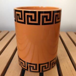 May include: Orange ceramic mug with a black Greek key design around the top and bottom.