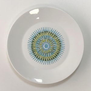 May include: A white ceramic plate featuring a central circular pattern composed of radiating triangular shapes in shades of light blue, olive green, and dark green, arranged in concentric rings around a solid light blue centre circle.