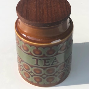 May include: A cylindrical ceramic tea canister featuring a glossy brown and olive green geometric pattern. The word TEA is embossed in the centre. The container is topped with a circular, dark brown wooden lid with a visible wood grain texture.