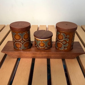 May include: A vintage salt and pepper set with a matching condiment jar. The ceramic containers are brown with a repeating green and blue floral design. The lids are made of wood. The set sits on a wooden tray.