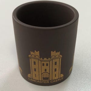 May include: A matte dark brown cylindrical mug featuring a gold-coloured graphic of a castle structure. The text "LANCASTER CASTLE" is printed in gold lettering at the base of the castle illustration. The mug has a smooth, solid-coloured exterior.