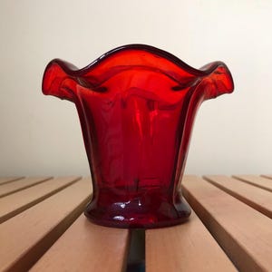 May include: A small, red glass vase with a ruffled rim. The vase is sitting on a wooden surface.