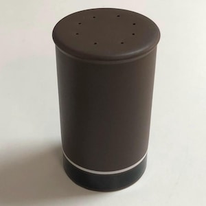 May include: A brown cylindrical salt shaker with a flat top featuring several small holes. The shaker has a black base with a white accent line. The design is simple and modern.