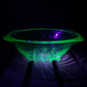 May include: A glowing green glass bowl with a scalloped rim and base. The bowl has two small handles and is illuminated with a black light, creating a vibrant neon effect. The bowl is likely vintage.