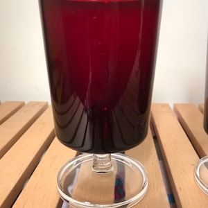 May include: Two ruby red wine glasses. Each glass features a cylindrical bowl and a clear, circular base. The short stem is transparent. The glasses are placed on a wooden surface.