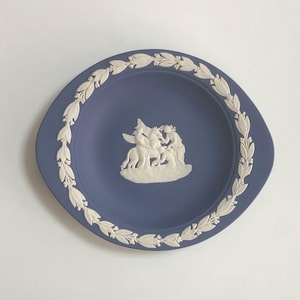 May include: A blue decorative plate with a white relief design. The plate features a central relief of figures and a border of white leaves. The plate has a unique shape with two handles. The plate is likely made of ceramic or porcelain.