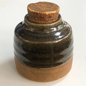 May include: A handmade ceramic jar with a dark brown glaze and a natural cork stopper. The jar has a rounded shape with a textured, ridged design. The base is a lighter, unglazed brown. A rustic, functional piece.