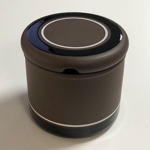 May include: A brown ceramic sugar bowl with a black and white lid. The lid has a circle design with a white outline and a black centre.