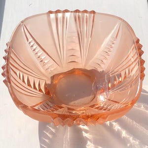 May include: A vintage, square pink glass bowl. The bowl has an etched design with geometric patterns and a scalloped rim. The bowl is likely for serving or display. The dimensions are in centimetres.
