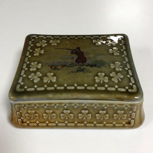 May include: A square, green ceramic trinket box with a raised shamrock design. The lid features a scene of a hunter with a dog in a field.