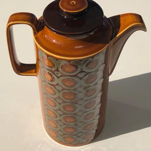 May include: A tall ceramic coffee pot featuring a glossy amber-brown glaze with a dark brown lid and handle. The cylindrical body is decorated with a repeating pattern of interlocking olive-green and orange circles that wrap around the centre.